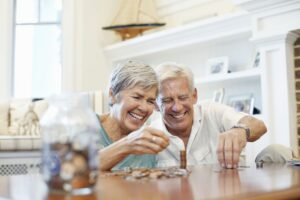 Couple of smiling retirees counting coins at home while organizing savings, illustrating financial planning and diversification strategies for retirement in the US.