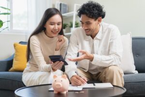 Couple reviewing expenses and planning their household finances in the US using a calculator and notebook while sitting on the living room sofa with a piggy bank nearby.